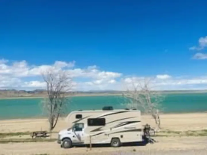 This **rv rental** is perfectly parked by a stunning turquoise lake with bare trees, ideal for a scenic road trip.. Winnebago Minnie Winnie 2020