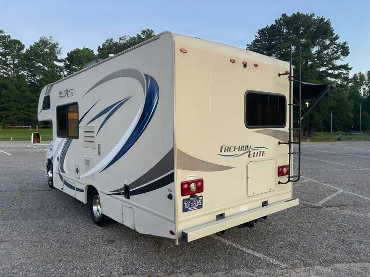 A beige Freedom Elite Class C rv rental, parked in an outdoor lot. Features a rear ladder and distinct side graphics, ready for travel.. Thor Freedom Elite 2018