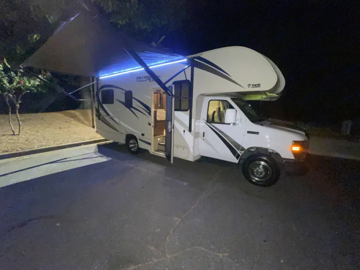 White Thor Freedom Elite RV rental with bright blue awning lights, parked at night with door open. Perfect for travel.. Thor Freedom Elite 2018