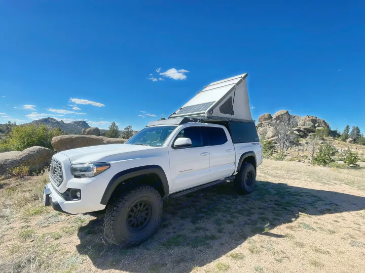 White Toyota Tacoma RV rental with pop-top camper & solar panels, parked in a rugged mountain setting.. 2023