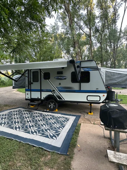 An RV rental camper with an extended awning and outdoor rug is set up at a peaceful, tree-shaded campsite.. Jayco Jay Feather 2018