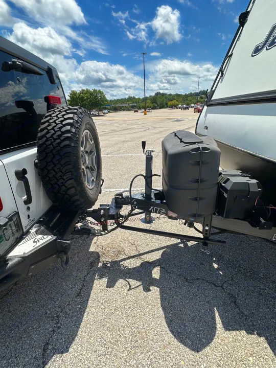 A white Jeep is hitched to an RV rental travel trailer in a sunny parking lot, showing the towing setup ready for travel and camping.. Jayco Jay Feather 2018
