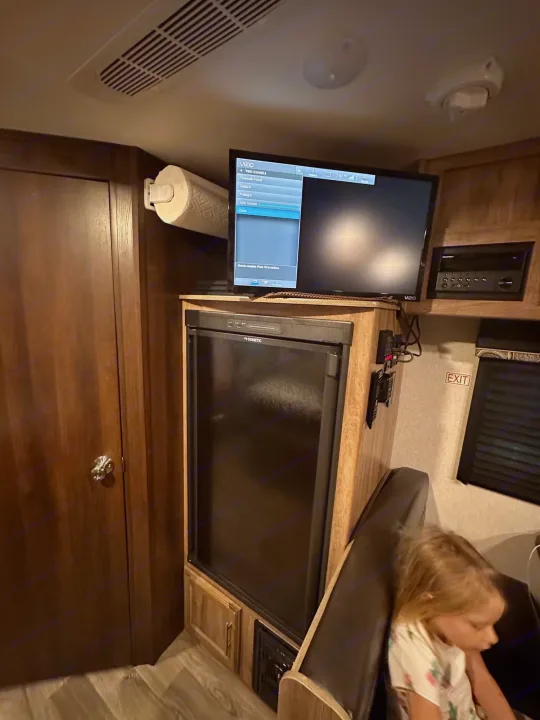 Inside an RV rental, showing a compact living space with a TV, refrigerator, and a child seated.. Jayco Jay Feather 2018