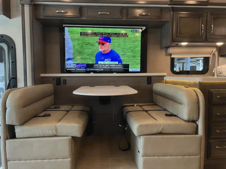 RV rental interior: a comfy dinette with a large screen TV showing an NFL game. Enjoy entertainment on your journey.. Entegra Coach Vision XL 2023