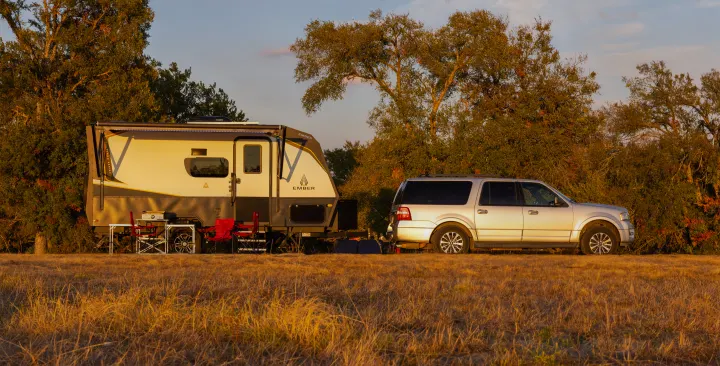 An RV rental setup: A silver SUV with a white Ember travel trailer, parked in a sunlit field surrounded by trees. Perfect for outdoor travel!. Ember Overland 2022