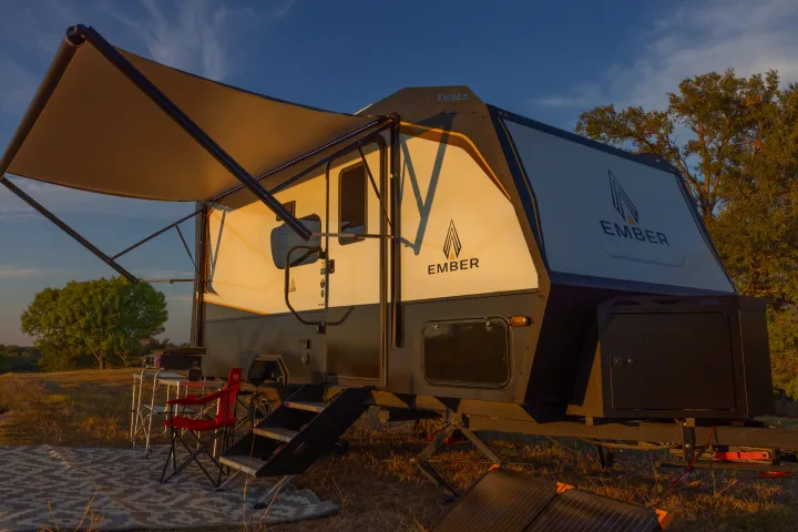 An Ember RV rental is set up for outdoor living with an extended awning, camping chairs, and ground solar panels at sunset.. Ember Overland 2022
