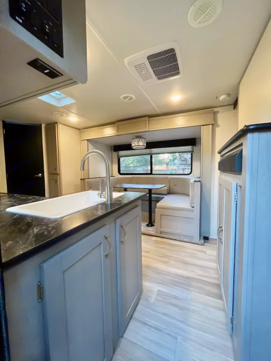 This RV rental interior features a modern kitchen with a large sink, light wood flooring, and a cozy dinette with window views.. Forest River Tracer 2022