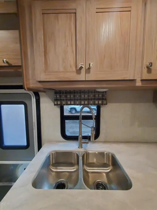 RV rental kitchen: light wood cabinets, double stainless steel sink, and modern faucet. A white Jeep is visible through the window.. Forest River Sunseeker 2019