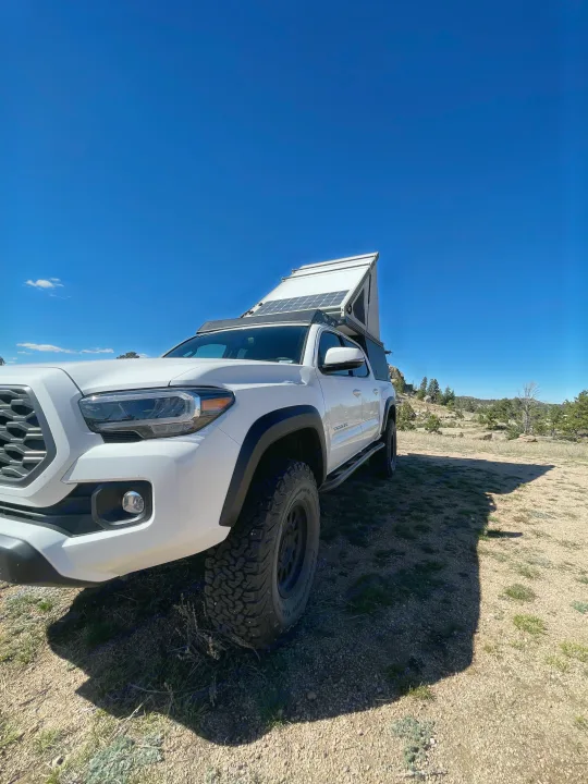 White Toyota Tacoma overlander with pop-top camper & solar panels, parked on a dirt path under clear blue sky. Ideal for RV rental trips.. Toyota Tacoma TRD Off-Road 2023