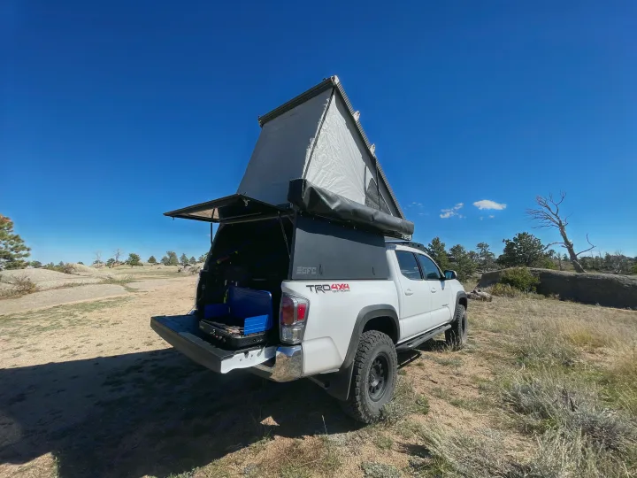 This white Toyota Tacoma with an open GFC pop-up camper is set up for outdoor cooking in a rocky landscape, perfect for your next rv rental.. Toyota Tacoma TRD Off-Road 2023