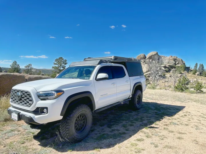 A white Toyota Tacoma truck with an overland camper setup, parked in a sunny, rocky landscape. Perfect for your next RV rental adventure.. Toyota Tacoma TRD Off-Road 2023