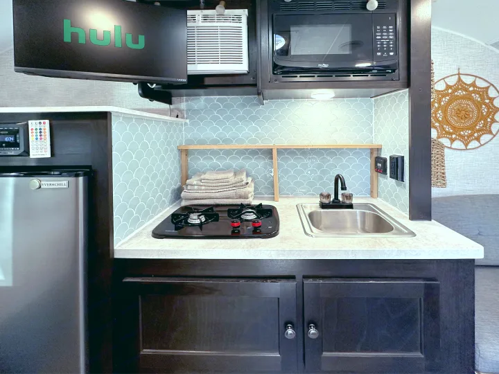 This RV rental kitchen features a sink, stovetop, microwave, fridge, TV, AC, and blue scallop tile backsplash.. Braxton Creek Bushwhacker Plus 2022
