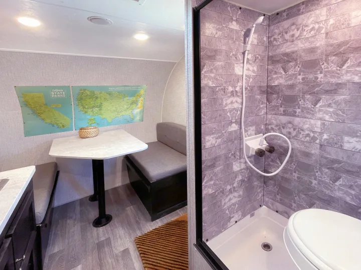 RV rental interior: dining nook with maps for travel planning, beside a compact shower and toilet with elegant grey tiles.. Braxton Creek Bushwhacker Plus 2022