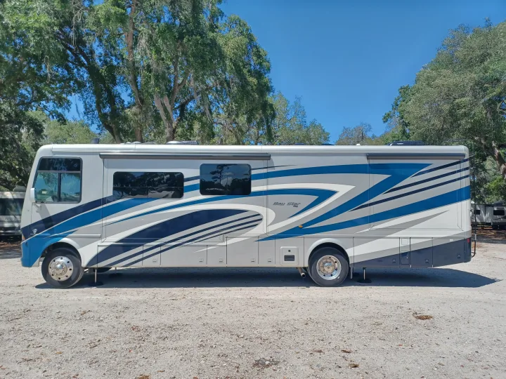 Grey & blue Bay Star Class A RV rental parked on a sunny gravel lot, surrounded by trees. Ideal for outdoor adventures.. Newmar Bay star 2022