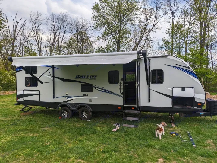 A modern Bullet Ultra Lite RV rental is set up on a grassy campsite with trees. Two small dogs enjoy the outdoor space.. Keystone RV Bullet Ultra Lite 2018
