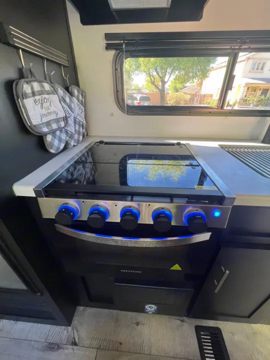 Inside a modern RV rental, featuring a sleek kitchen with an illuminated Greystone stove, oven, and window view.. Keystone RV Springdale 2022