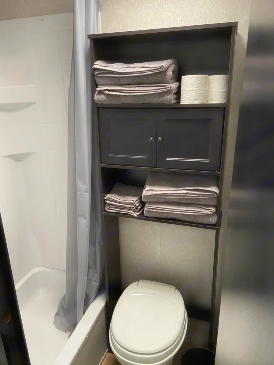 Compact RV rental bathroom featuring a toilet, shower, and dark shelving unit with fresh towels and toilet paper.. Cherokee wolf pup 2021