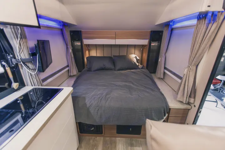 Inside a modern RV rental: a spacious gray bed, compact kitchen with cooktop, wall-mounted TV, and blue ambient lighting. Cozy travel.. Black Series Black Series Trailer 2022
