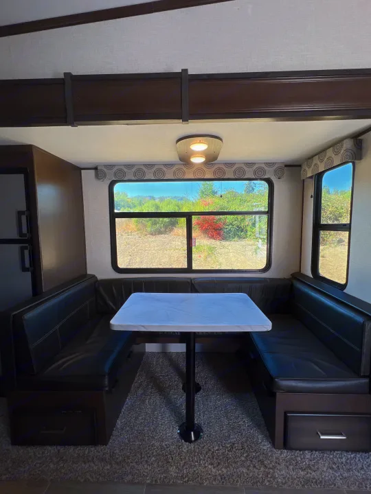 RV rental dinette featuring dark U-shaped bench seating, a white table, and large windows showing outdoor scenery.. Atlas Atlas Fifth Wheel 2020