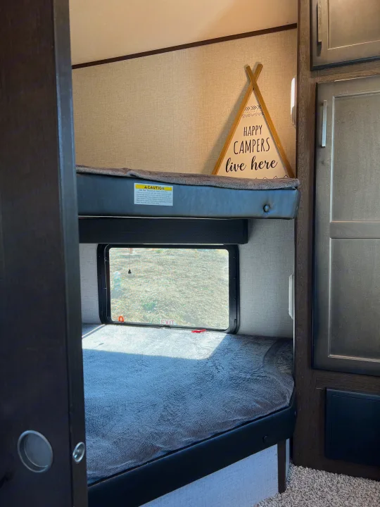 Cozy RV rental bunk beds, offering a comfortable sleeping area with a window view and "Happy Campers" sign.. Atlas Atlas Fifth Wheel 2020