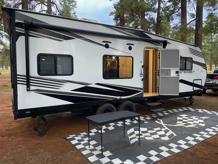 This black and white RV rental is set up with an awning, table, and checkered mat at a forest campsite.. Genesis Supreme Rv Genesis Supreme 2022