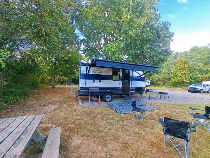 Blue and white rv rental, "Wolf Den" travel trailer, parked at a spacious campsite with an extended awning, chairs, and picnic table.. Forest River Cherokee 2024