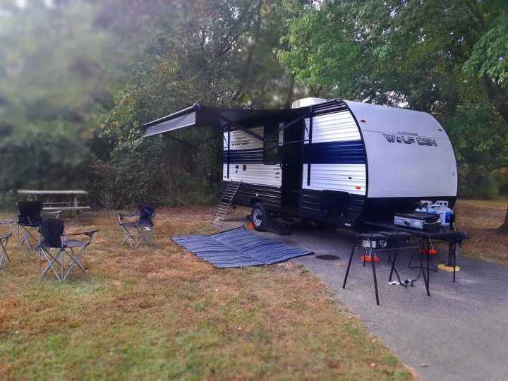 This RV rental, a Cherokee Wolf Den travel trailer, is set up at a spacious campsite with chairs, rug, and grill.. Forest River Cherokee 2024