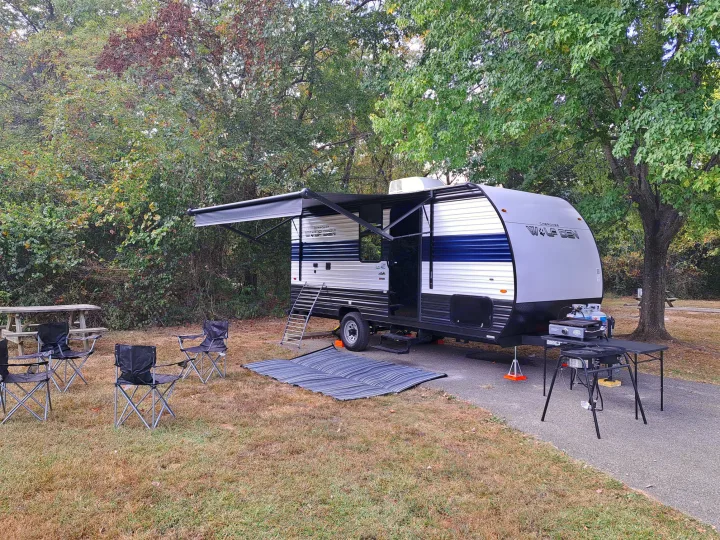 RV rental campsite setup featuring a travel trailer with extended awning, outdoor chairs, grill, and picnic table for camping.. Forest River Cherokee 2024