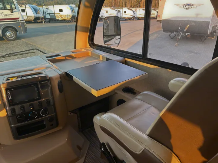 View from an RV rental cockpit, showing the dashboard, passenger seat, a pull-out table, and other RVs parked outside.. Thor Axis 2018