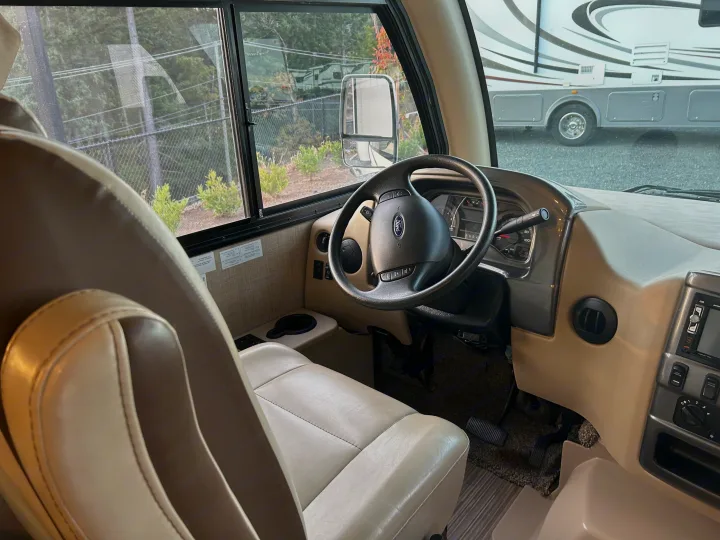 Spacious RV rental interior: beige driver's seat, Ford steering wheel, dashboard, and outside view of another RV.. Thor Axis 2018