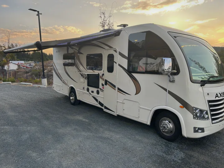 Cream-colored rv rental with extended awning, glowing LED lights, and a tall pole, parked on a gravel lot at sunset for travel.. Thor Axis 2018