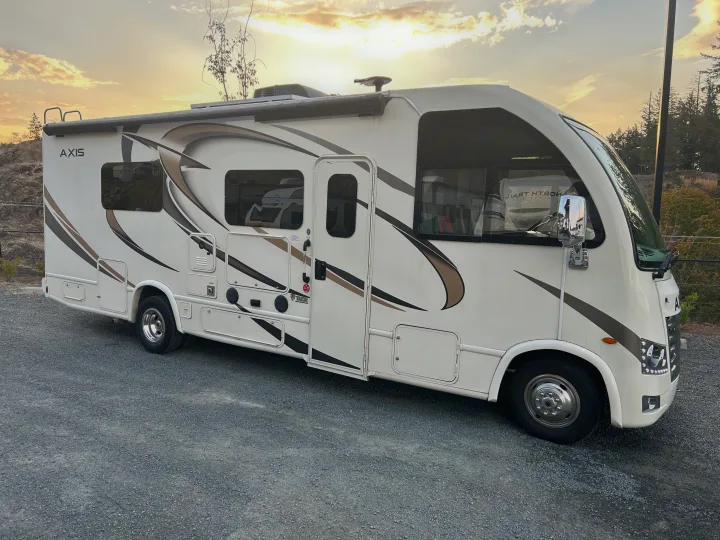 Side view of a white Thor Axis RV rental with brown and black decals, parked on gravel, set against a sunset sky.. Thor Axis 2018