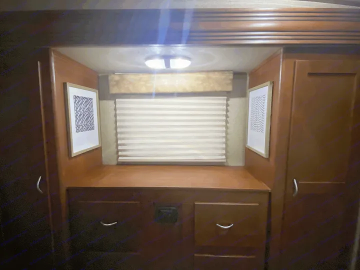 Cozy RV rental bedroom vanity with rich wood cabinets, a window with blinds, framed art, and overhead lighting.. Forest River Vengeance 2015