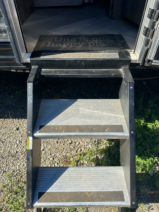 RV rental entrance with black and silver steps extended to gravel ground. A doormat and caution labels are visible inside.. Forest River Salem Cruise Lite 2021