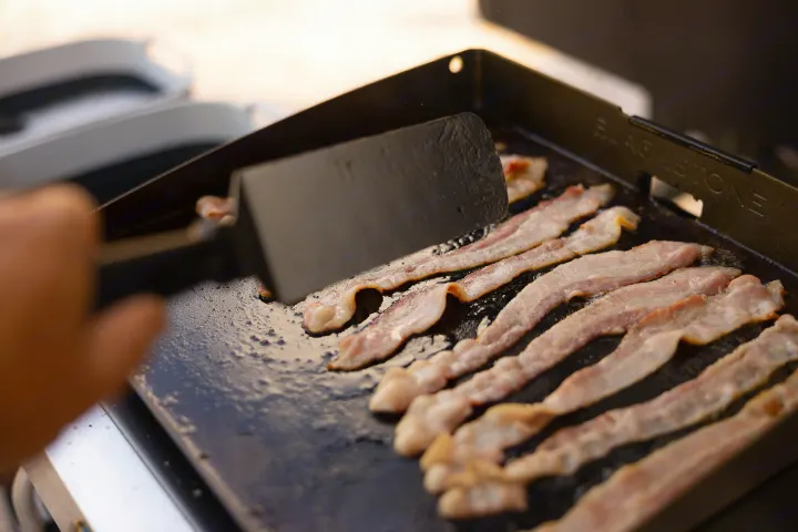 A hand cooks sizzling bacon on a Blackstone griddle. Perfect for your RV rental getaway and outdoor breakfast.. Ember Overland 2022