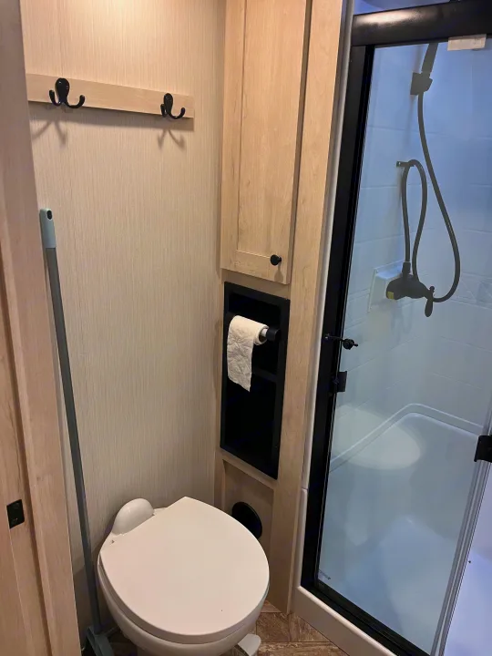 This RV rental features a compact, light-toned bathroom with a toilet, recessed toilet paper holder, and a clear glass shower stall.. Forest River Wildcat 2023