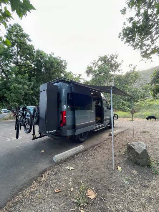 Gray Sandy Vans RV rental parked with awning out, bikes on back, ready for adventure. Dog nearby.. Mercedes-Benz Sprinter RV Motorhome Campervan 2020