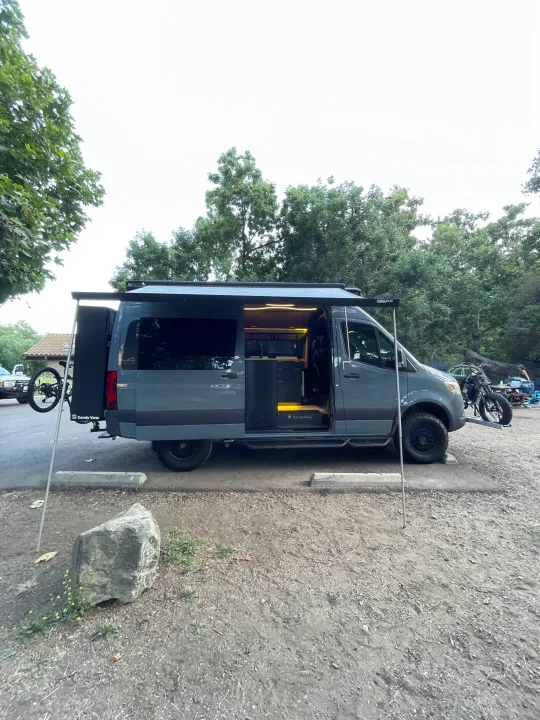A customized Sandy Vans RV rental parked in nature. This dark grey camper van features an extended awning, bike rack, and lit interior.. Mercedes-Benz Sprinter RV Motorhome Campervan 2020