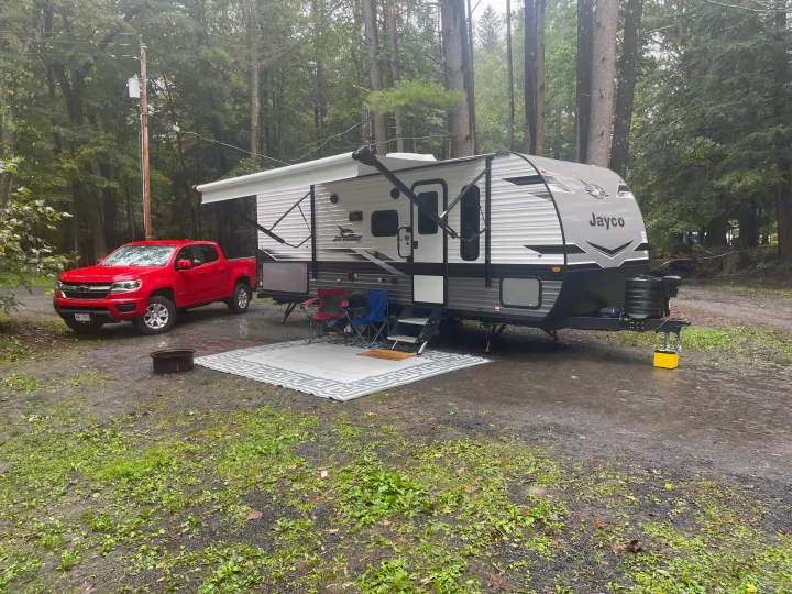 A grey Jayco travel trailer RV rental with awning, red pickup, and chairs at a muddy, wooded campsite.. Jayco Jay Flight 2023