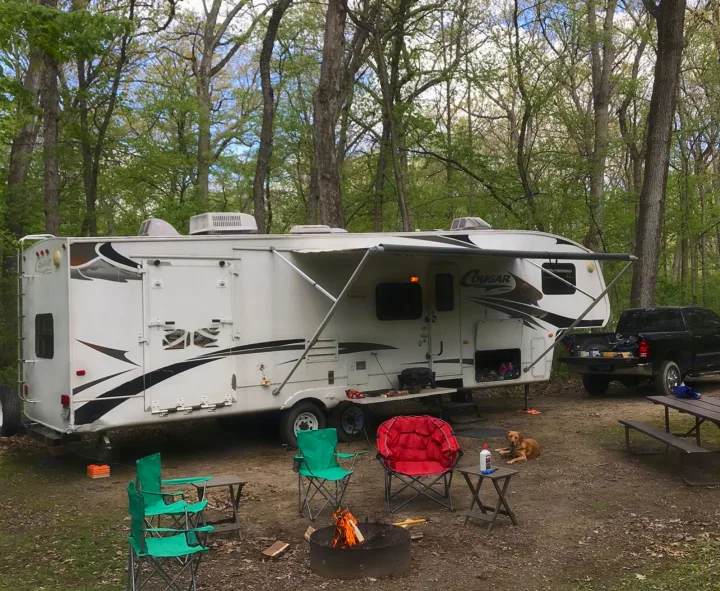 An RV rental and truck are parked at a wooded campsite, featuring a campfire, chairs, and a dog.. Keystone RV Cougar 2012