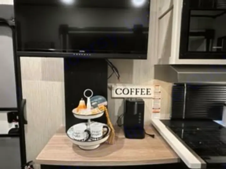 This compact RV rental kitchen features a coffee station: a two-tiered tray, mugs, coffee maker, and "COFFEE" sign under a TV.. Forest River Wildcat 2022