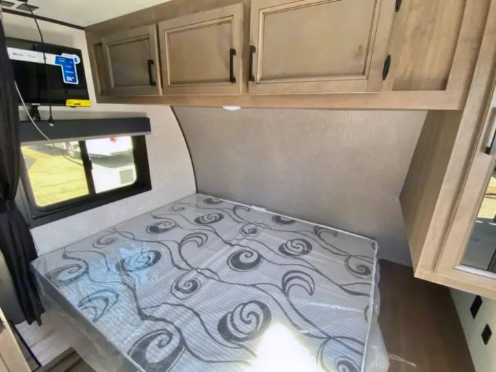 RV rental bedroom with a patterned mattress, overhead cabinets, mounted TV, and window, ready for your next adventure.. Starcraft Autumn Ridge 2023