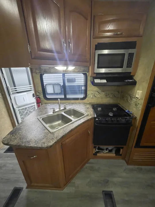 Inside an RV rental, showing a functional kitchen with wood cabinets, a double sink, microwave, and gas stove.. Keystone RV Cougar 2012