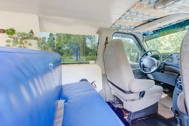 Converted van interior for RV rental: cozy blue seating, driver's cockpit, and serene park view from side window.. Ford Ford E350 2007
