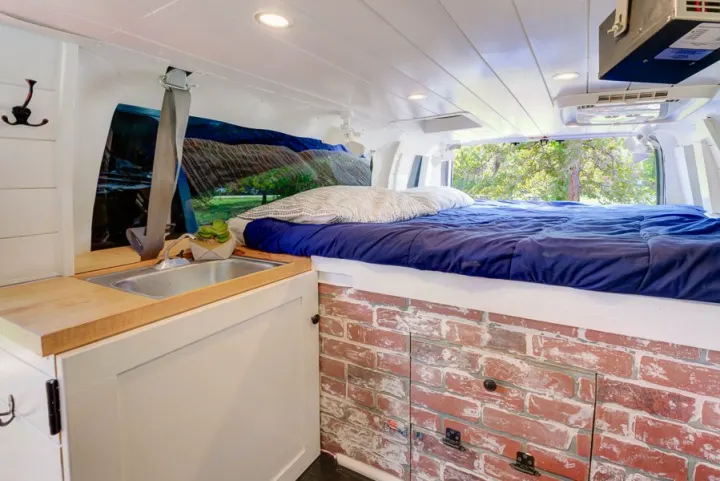 Inside a custom rv rental, a cozy bed with blue bedding rests on brick storage next to a compact kitchen with a sink.. Ford Ford E350 2007
