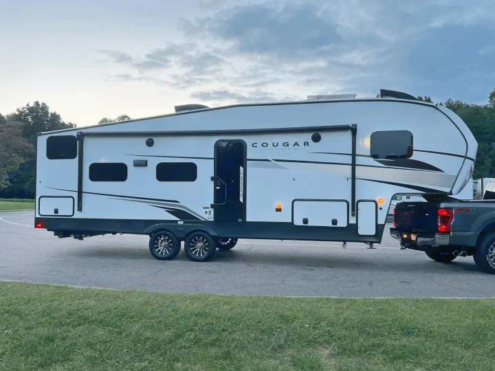 Hooked up for travel: a sleek white fifth-wheel RV, perfect for an RV rental, towed by a pickup truck against a cloudy sky.. Keystone Cougar Sport 2024