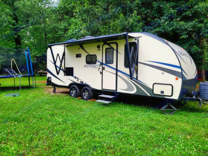 A beige Sonic travel trailer (RV) with awning out, parked on lush grass amidst trees and a trampoline. Ideal for an RV rental getaway.. Sonic Sonic Trailer 2019