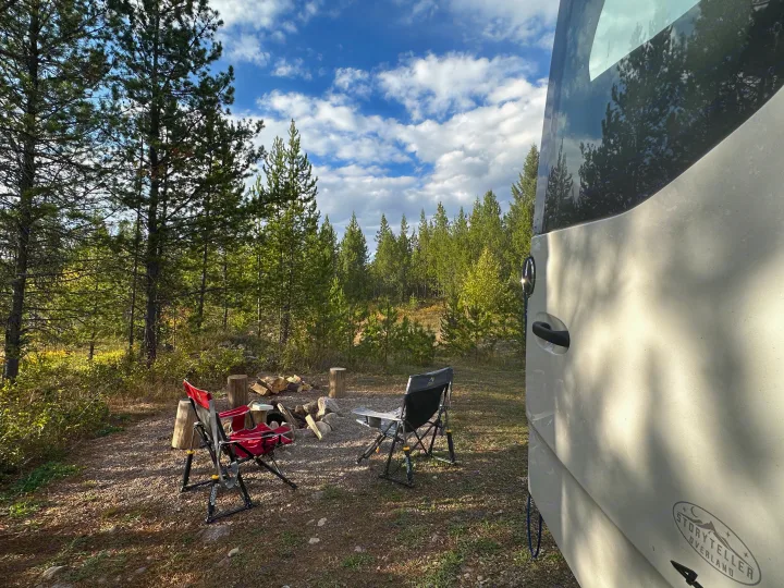 RV rental at a serene forest campsite, featuring camping chairs around a fire pit amidst tall pine trees under a blue sky.. Storyteller Overland Storyteller Overland Class B 2020