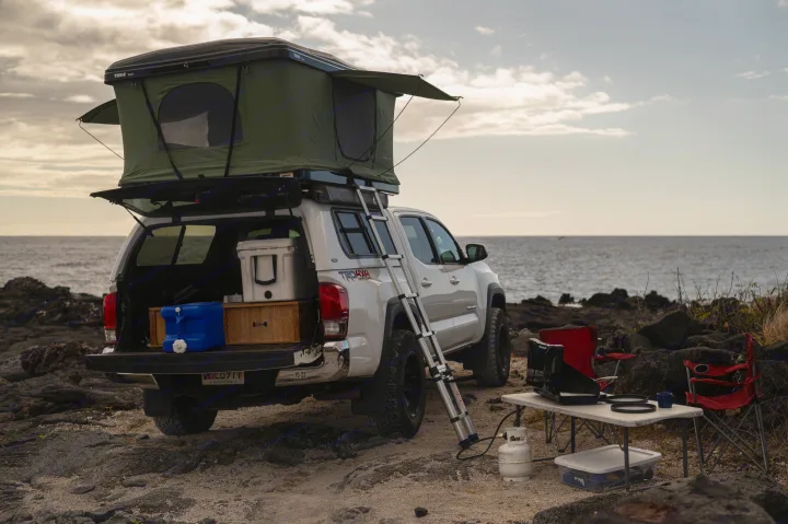 Explore with this RV rental: White Toyota pickup with rooftop tent, ladder, and full camp setup on a rocky beach.. Custom Camper 2016