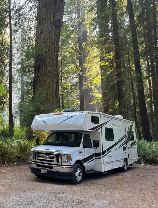 White RV rental parked on a dirt road in a majestic redwood forest, with sunlight filtering through tall trees.. Forest River Coachmen Freelander 2021